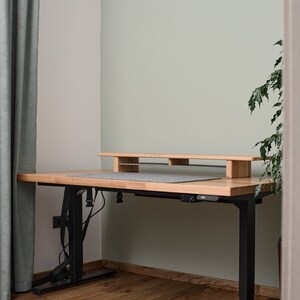 May include: A wooden standing desk with a black metal frame and a gray desk mat. The desk has a built-in shelf for a monitor or other items. The desk is in a room with light green walls and wooden floors.