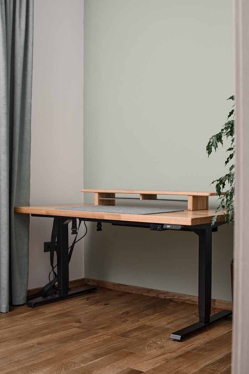 May include: A wooden standing desk with a black metal frame and a gray desk mat. The desk has a built-in shelf for a monitor or other items. The desk is in a room with light green walls and wooden floors.
