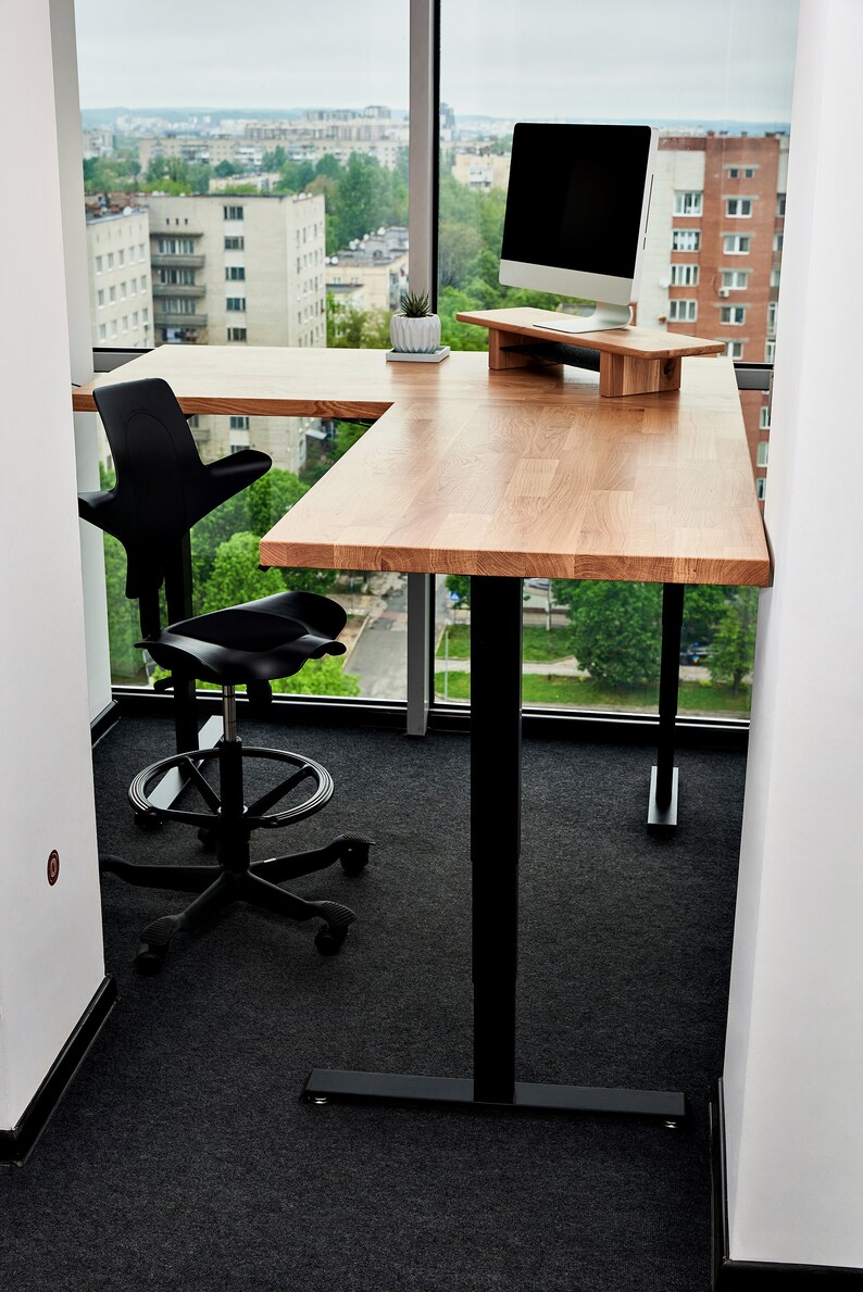 May include: A black office chair with a unique design sits next to a wooden desk with a black metal frame. The desk has a monitor stand and a potted plant on the surface. The desk is in front of a large window with a view of a city skyline.