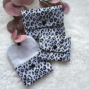 Könnte beinhalten: Drei kleine, graue und schwarze Stoffbeutel mit Leopardenmuster und Knopfverschluss. Die Beutel sind in verschiedenen Größen und auf einer weißen, flauschigen Oberfläche angeordnet.