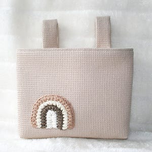Könnte beinhalten: Eine beige Fahrradlenkertasche aus Waffelstoff mit einem gehäkelten Regenbogen-Applikationen.