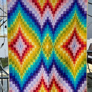 May include: A vibrant, handmade quilt with a geometric design. The quilt features a repeating pattern of colorful squares in a rainbow of hues, including red, orange, yellow, green, blue, and purple. The quilt is displayed outdoors, showcasing its bright colors.