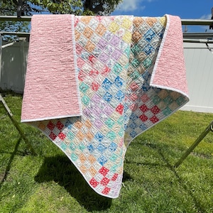 May include: A colorful patchwork quilt with a pink backing and white trim, hanging on a clothesline. The quilt features a variety of square patterns in pastel colors, including blue, yellow, red, and green. The quilt is made of fabric.