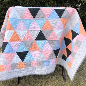 May include: A quilt with a geometric pattern of triangles in shades of pink, blue, gray, and black. The quilt is made with a white background and has a border of gray and peach.