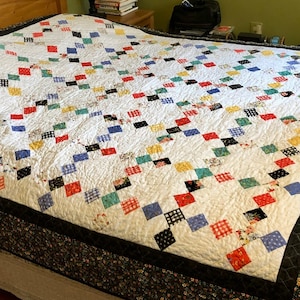 May include: A white quilt with a black, red, blue, yellow, and green patchwork design. The quilt has a black border with a floral pattern.