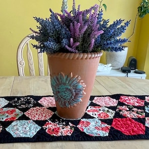 May include: A decorative table setting featuring a terracotta pot with a turquoise sunflower design, filled with purple and blue faux lavender. A floral patterned table runner with black background and red, pink, and blue floral designs.