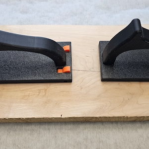 May include: Two black sanding blocks with orange handles on a wooden surface.