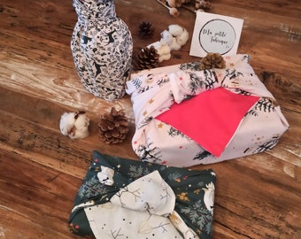 May include: Two gifts wrapped in fabric with a festive winter design. One gift is a bottle wrapped in a white fabric with black and white deer and snowflakes. The other gift is a box wrapped in a pink fabric with gold stars and a white fabric with a green and white winter design. The gifts are on a wooden surface with dried flowers and cotton balls.