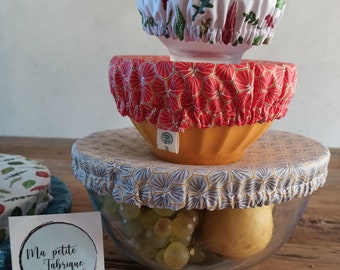 Puede incluir: Tres cubiertas de alimentos reutilizables hechas de tela con diferentes patrones. Las cubiertas están apiladas una encima de la otra, con la cubierta superior siendo blanca con diseños florales verdes y rojos, la cubierta del medio siendo roja con diseños hexagonales amarillos y la cubierta inferior siendo blanca con diseños hexagonales azules y amarillos. Las cubiertas están sobre un tazón de vidrio lleno de uvas y una pera. El tazón está sentado sobre una mesa de madera. Una pequeña tarjeta blanca con el texto "Ma petite Fabrique" está en primer plano.