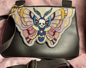 Può includere: Borsa a tracolla nera con un design a falena ricamato. La falena presenta un teschio e ali nei toni del rosa, giallo, blu e viola. La borsa ha una tracolla regolabile nera e finiture color argento.