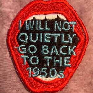Könnte beinhalten: Ein gestickter Aufnäher in Form eines offenen Mundes mit roten Lippen und weißen Zähnen. Das Innere ist schwarz mit hellblau gesticktem Text: I WILL NOT QUIETLY GO BACK TO THE 1950s. Der Aufnäher hat einen dicken roten Rand und detaillierte Stickerei.