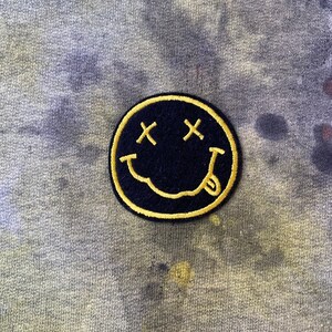 May include: A yellow embroidered smiley face patch with black eyes and a mouth, on a gray and yellow tie-dye fabric.