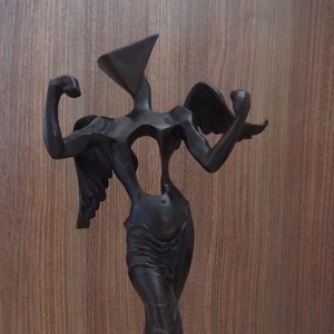 May include: A black bronze sculpture of a figure with wings and a geometric head. The figure is standing on a black base.