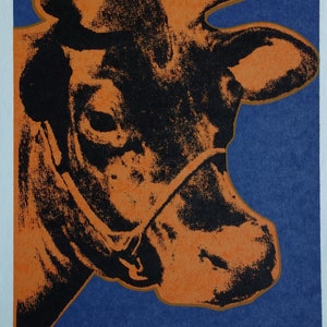May include: A close-up of a black and orange cow print. The cow's head is turned to the side, and its eye is visible. The background is a solid blue color. The print is signed "Andy Warhol" in the bottom right corner.
