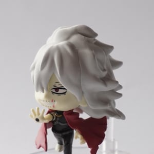 May include: A small figurine of a character with white hair and a red cape. The figure has a black outfit and a pale complexion with visible scars. It stands on a clear, hexagonal base.