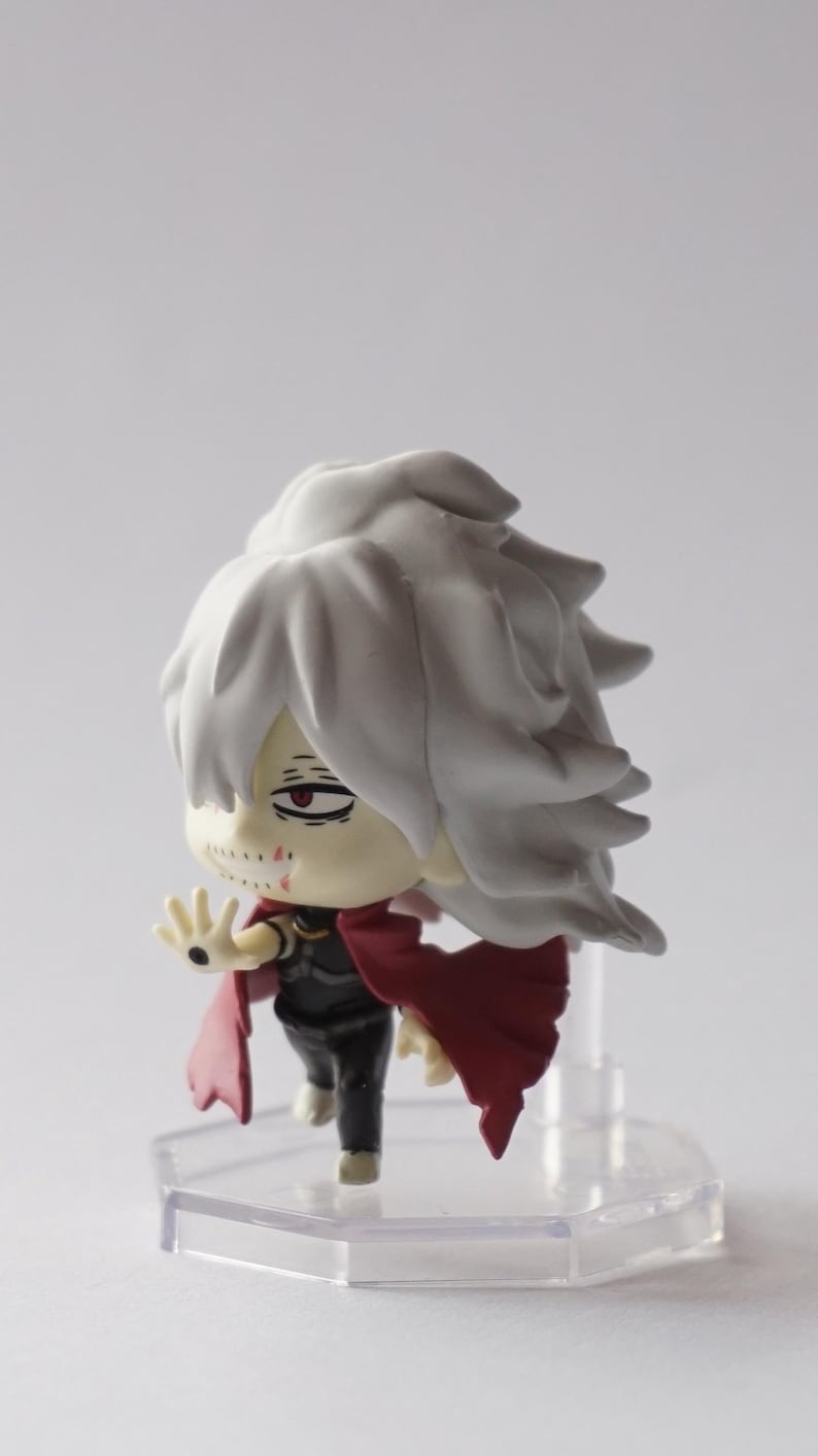 May include: A small figurine of a character with white hair and a red cape. The figure has a black outfit and a pale complexion with visible scars. It stands on a clear, hexagonal base.