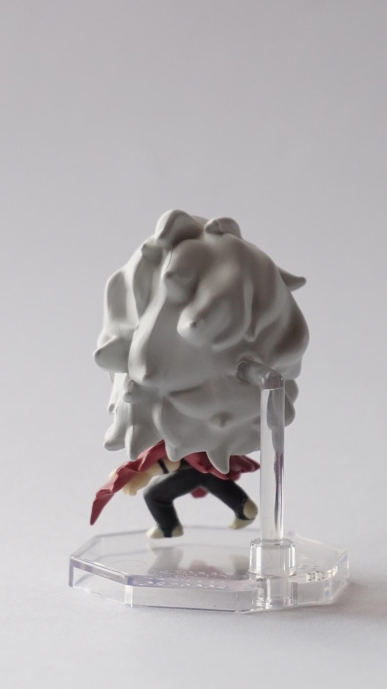 May include: A small figurine of a character with a large, white, cloud-like hairpiece. The figure is wearing a red garment and black pants. It is mounted on a clear plastic stand.