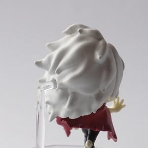 May include: A small figurine of a character with white spiky hair and a red cape. The figure is mounted on a clear plastic stand. The figure is viewed from the back, with the character's arm raised.