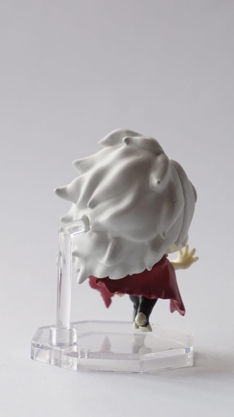 May include: A small figurine of a character with white spiky hair and a red cape. The figure is mounted on a clear plastic stand. The figure is viewed from the back, with the character's arm raised.