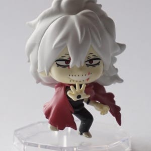 May include: A small figurine of a character with white hair and a red cape. The figure has a menacing expression with red eyes and a stitched-up mouth. It stands on a clear, hexagonal base.