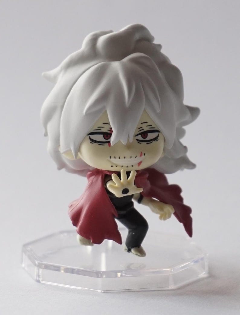 May include: A small figurine of a character with white hair and a red cape. The figure has a menacing expression with red eyes and a stitched-up mouth. It stands on a clear, hexagonal base.