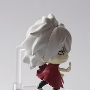 May include: A small figurine of a character with white hair and a red cape. The figure is on a clear plastic stand. The character has a determined expression and is in a dynamic pose, with one arm raised.