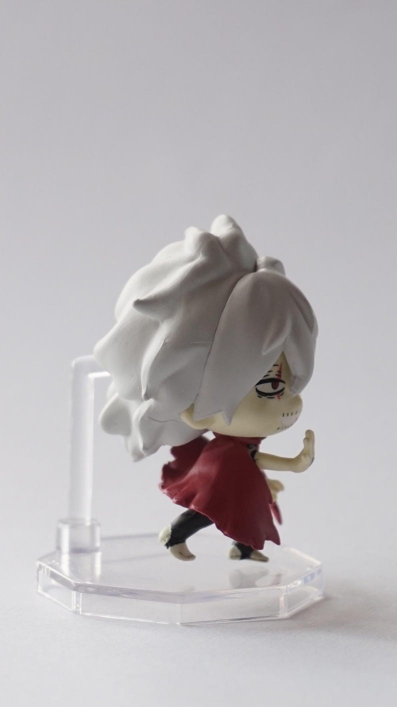 May include: A small figurine of a character with white hair and a red cape. The figure is on a clear plastic stand. The character has a determined expression and is in a dynamic pose, with one arm raised.
