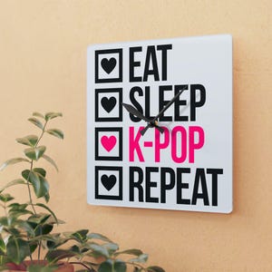 May include: White square clock with black and pink text that reads "Eat Sleep K-Pop Repeat" with a black clock hand.