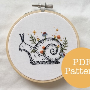 Könnte beinhalten: Ein gestickter Schnecken mit Blumen und Sternen auf einem weißen Stoff in einem hölzernen Stickrahmen. Der Rahmen liegt auf einer weißen Oberfläche. Der Text "PDF Pattern" befindet sich auf einem gelben Kreis.