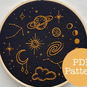 Pode incluir: Um aro de bordado preto com um desenho de cena celestial em fio dourado. O desenho inclui um sol, uma lua, estrelas, planetas e uma galáxia. O texto "PDF Pattern" está em um círculo amarelo no canto inferior direito.