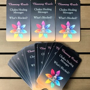 K&ouml;nnte beinhalten: Ein Kartendeck mit rechteckigen Orakelkarten, die den Text Charming Oracle, Chakra Healing Messages und What's Blocked? zeigen. Jede Karte zeigt eine zentrale Figur in sitzender Meditationshaltung, umgeben von einem bunten Bl&uuml;tenmotiv.