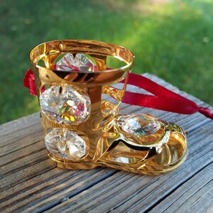 May include: A gold-colored decorative boot with clear, sparkling crystals. The boot has a red ribbon attached, suggesting it may be an ornament. The boot is sitting on a weathered wooden surface, with a blurred green background.