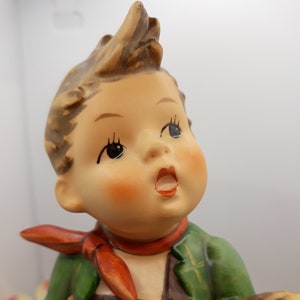 May include: A porcelain figurine of a boy wearing a green jacket, brown lederhosen, and a red scarf. He is holding a stick in his right hand and has a surprised expression on his face.