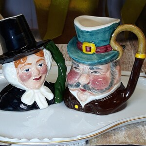 May include: Two ceramic mugs shaped like figures, one with a black top hat and the other with a green hat and a pipe-shaped handle. The mugs are on a white tray with gold trim. The figures have painted faces.