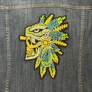 May include: A colorful embroidered patch featuring a skull with a headdress of green and yellow feathers. The skull is white with black details and a yellow mouth.