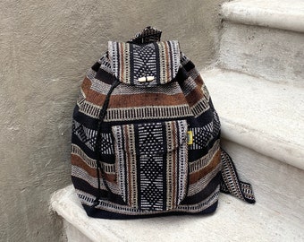May include: A woven backpack with a geometric pattern in black, brown, and white. The bag has a flap closure with wooden toggles and a small front pocket. The backpack is sitting on a concrete surface.