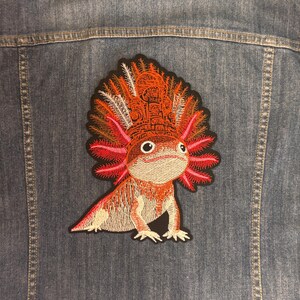 May include: An embroidered patch featuring an axolotl with a large, ornate, orange and red headdress, attached to the back of a blue denim jacket. The axolotl has pink gills, a light tan body, and a black border surrounding the entire design.