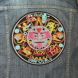 Puede incluir: Un parche redondo bordado con un diseño de calendario azteca colorido. El parche es principalmente negro con ilustraciones detalladas en tonos de rosa, naranja, amarillo y turquesa. Está adherido a una chaqueta vaquera.