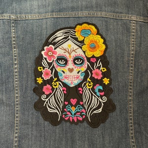 May include: Embroidered iron-on patch featuring a colorful Day of the Dead sugar skull with floral accents. The skull is depicted as a woman with black hair, blue eyes, and a pink, white, and blue painted face.