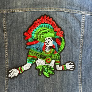 May include: A green, red, and blue sequined patch of a skeleton figure wearing a headdress on a denim jacket.
