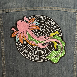 Pode incluir: Patch bordado com um axolotl rosa com detalhes em laranja, azul e verde, sobre um fundo circular preto com símbolos brancos. O patch está preso a uma jaqueta jeans.