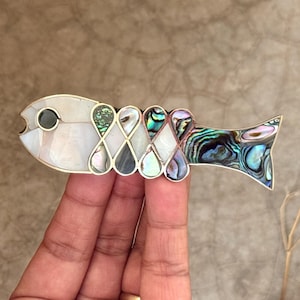 Może przedstawiać: Broszka w kształcie ryby z obrysem z metalu w kolorze srebrnym. Korpus wykonany z iryzującej muszli abalonu i masy perłowej, z czarnym okiem. Unikalny dodatek.