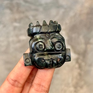May include: A dark green stone carving shaped like a stylized face with large circular eyes, a prominent brow, and a crown-like top. The object features small circular perforations on the left and right sides, showcasing a smooth, polished finish.