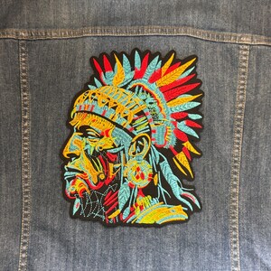 May include: A denim jacket with a large, colorful embroidered patch of a Native American chief. The patch features vibrant hues of yellow, red, blue, and green, depicting the chief in profile. The jacket is a classic blue wash.