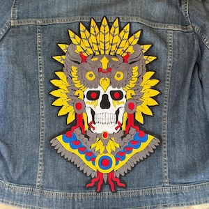 May include: A colorful embroidered patch featuring a skull with a feathered headdress. The skull is adorned with red, blue, and yellow details, and the headdress is made of yellow feathers.