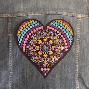 Op de afbeelding: Een hartvormige geborduurde patch met een zwarte rand, met een kleurrijk mandala-ontwerp. De mandala bevat blauwe, roze, gele en paarse stippen en bloempatronen. De patch is bevestigd aan een spijkerjasje.