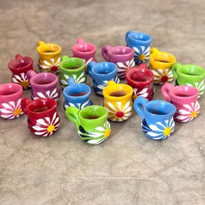 May include: A collection of miniature ceramic mugs in various bright colors including red, pink, yellow, green, and blue. Each small mug features a hand-painted white daisy pattern with a contrasting center dot on the exterior surface.