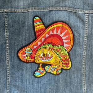 May include: A colorful embroidered patch of a smiling taco wearing a sombrero. The taco is holding a maraca in its hand. The patch is perfect for adding a fun and festive touch to any denim jacket, backpack, or other fabric item.