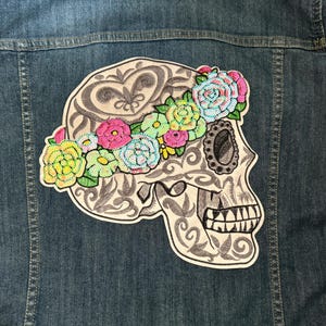 May include: A white embroidered skull with a floral crown of pink, yellow, and blue flowers on a denim jacket.