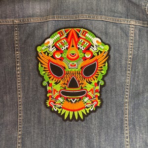 Puede incluir: Una chaqueta de mezclilla azul con un parche bordado grande e intrincado en la parte posterior. El parche muestra un diseño de máscara estilizado y simétrico en tonos vibrantes de naranja, verde lima, rojo y blanco sobre un fondo negro.